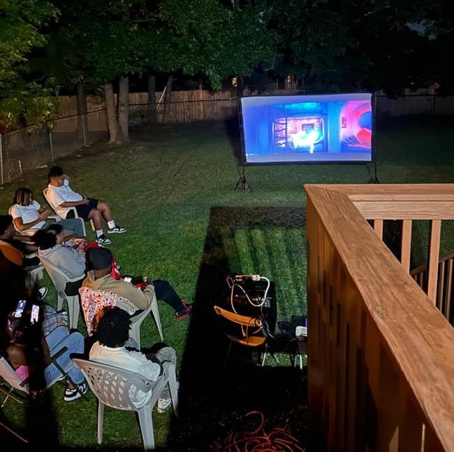 Outdoor movie nighttime setup with respective radical seated successful chairs watching a projected movie successful a backyard. Trees and a obstruction are visible
