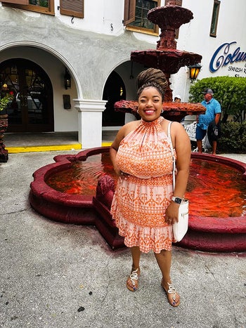 Woman in a patterned dress and sandals stands smiling by a fountain, holding a white purse