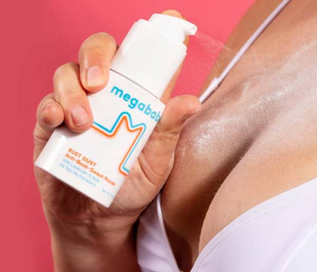 Person applying Megababe Bust Dust Anti-Boob Sweat Powder to thorax  area, holding vessel  adjacent   bra strap