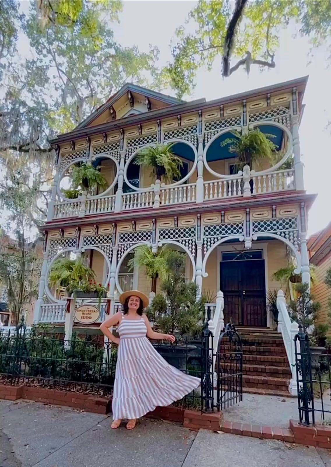 Person in a wide-brimmed hat poses in front of a Victorian-style house with intricate woodwork and ferns on the porch