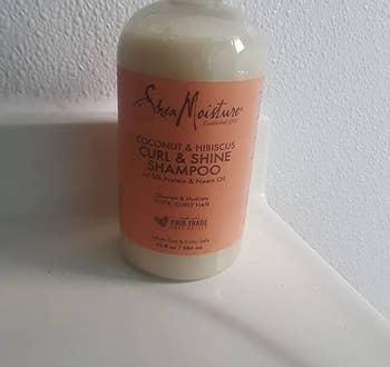 Reviewer image of white and peach colored curl shampoo bottle on top of white counter in font of a white wall