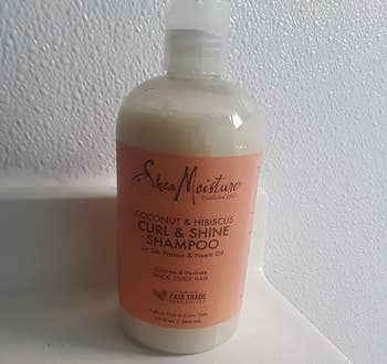 Reviewer image of white and peach colored curl shampoo bottle on top of white counter in font of a white wall