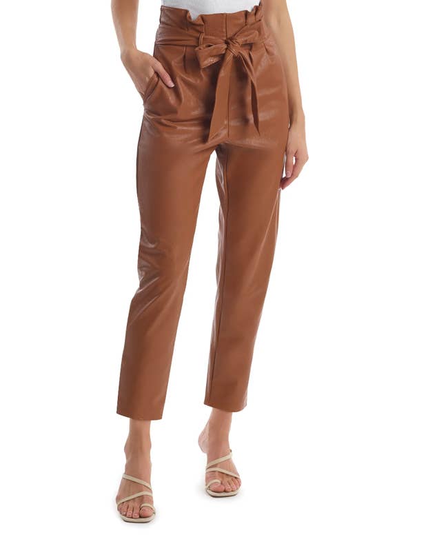 Model wearing brown straight fit faux leather paperbag pants with matching front tie in a bow, paired with nude strappy heels