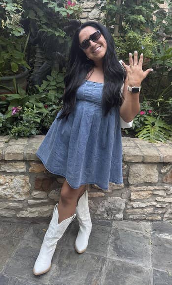 Person wearing a denim dress, achromatic boots, and sunglasses, smiling and showing a manus gesture, lasting successful a plot setting