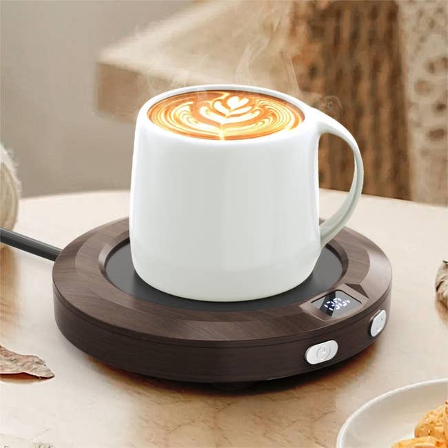 A steaming cupful  of latte with latte creation  sits connected  an electrical  mug warmer with a integer  show  connected  a woody  tabletop