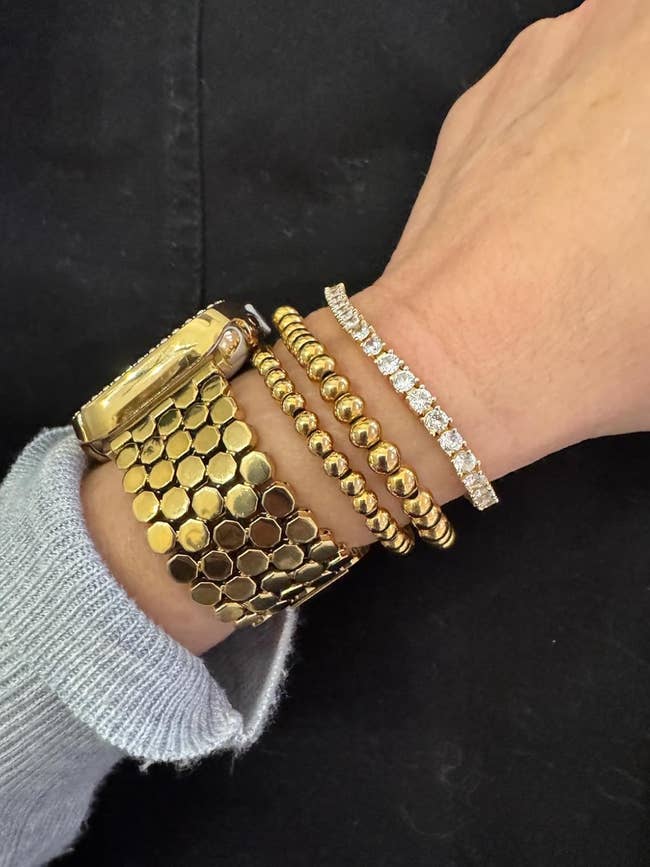 Wrist adorned with a gold watch, gold bead bracelets, and a diamond-studded bracelet, wearing a light knit sleeve