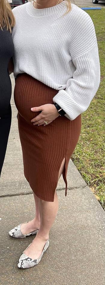 A idiosyncratic   wearing a achromatic  knit sweater, a ribbed brownish  skirt with a slit, and snake-print flats cradles their large   belly.