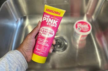 Person holds a tube of The Pink Stuff cleaning paste over a metal sink, highlighting product features like being vegan and effective on dirt