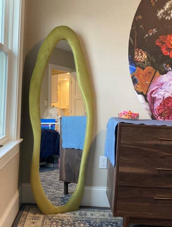 Full-length oval mirror beside a wooden dresser against a floral wallpapered wall in a room