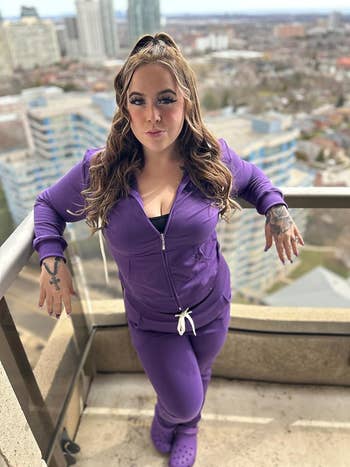Woman in a purple tracksuit posing on a balcony with cityscape in the background. She wears matching shoes and styled hair
