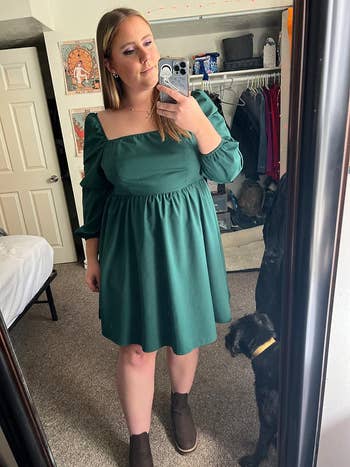 reviewer wearing a green dress with a square neckline and puffed sleeves, taking a mirror selfie in a bedroom; a small black dog is beside her