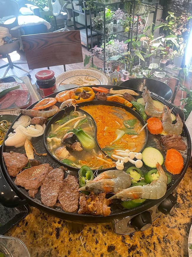 black electric grill with built-in hot pot heating up meats, veggies, and prawns