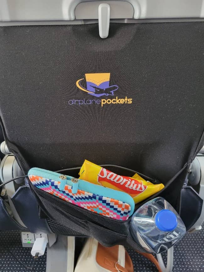 reviewer photo of the organizer on the back of an airplane seat holding a laptop case, a bottle of water, and a bag of chips