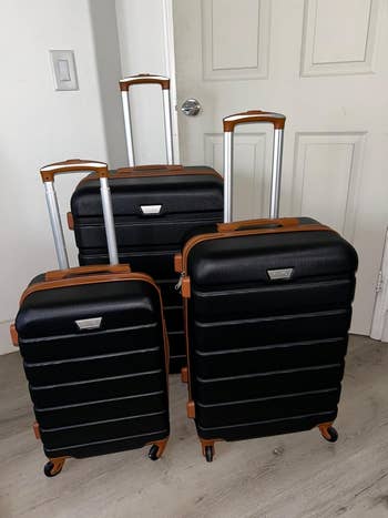 Three black, textured luggage pieces with metallic telescopic handles and brownish accents, lasting upright connected a wood floor