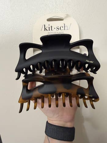 Two Kitsch hairsbreadth  claw clips made of recycled plastic, 1  achromatic  and 1  tortoiseshell, held successful  a manus  against a plain background
