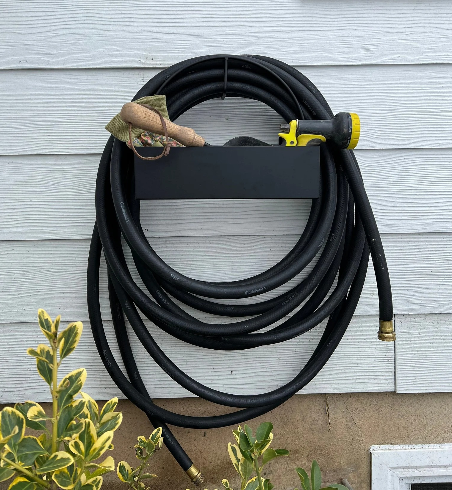 the black hose holder attached to the side of a house with a hose wrapped around it and various gardening supplies in its shelf