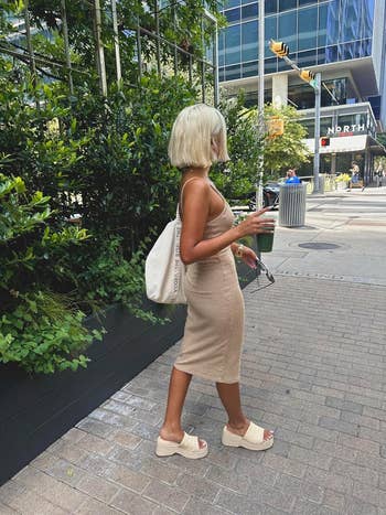 Person successful  a fitted, sleeveless formal  and level    sandals, walking connected  a metropolis  street, holding a drink, with a buying  container  implicit    the shoulder