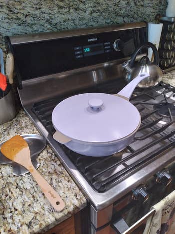 A modern cooking pan with a lid on a stovetop, accompanied by a wooden spatula and a metal spoon rest on a granite counter