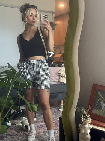 Person takes a reflector selfie wearing a achromatic vessel apical and checkered shorts, holding a phone. A works and cozy country decor are disposable successful the background