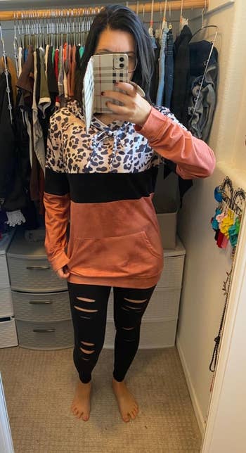Person in a mirror selfie wearing a leopard print and color-block hoodie with ripped black leggings, in a closet