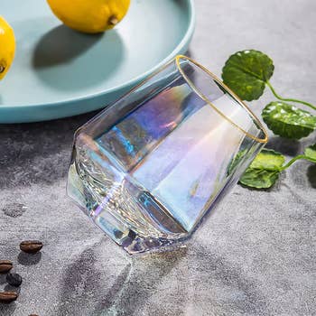 A tilted iridescent glass cup on a textured surface near a plate with lemons and coffee beans