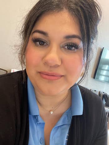 A reviewer in a casual outfit, wearing a black cardigan over a blue top, looks at the camera indoors