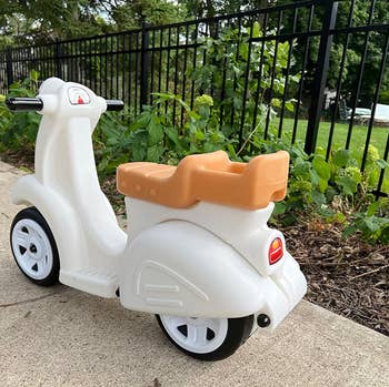 Child's ride-on toy scooter parked on a sidewalk near grass and a metal fence. Suitable for outdoor play