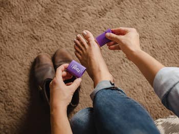 Person applying ft  balm with purple packaging connected  bare foot; brownish  boots successful  the inheritance  connected  carpet. Casual and comfy  setting