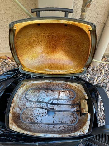 Outdoor electrical grill with lid open, showing a greasy interior and rusted heating element. The grill is acceptable connected a rocky ground
