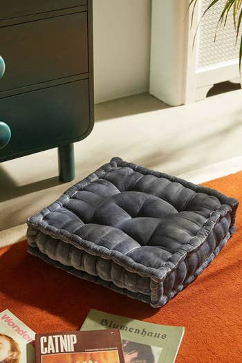 Plush, square floor cushion with tufted design on an orange rug, next to a magazine and furniture