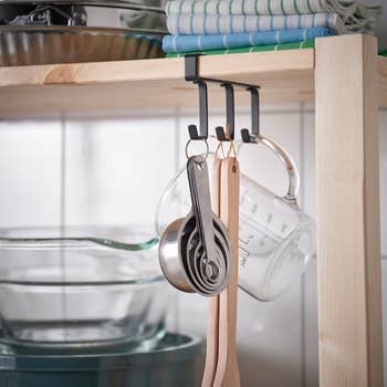 Measuring cups and wooden utensils hang from a shelf in a kitchen, next to stacked glass bakeware and dish towels