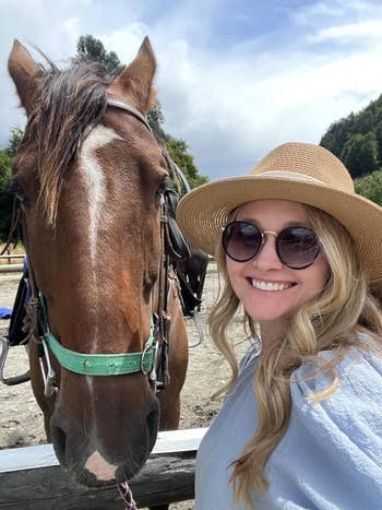reviewer in a straw hat and sunglasses smiling next to a horse in a bridle, outside during the day