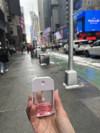 Hand holds Touchland Power Mist sanitizer connected  a engaged  metropolis  street, suggesting its convenience for usage  on-the-go