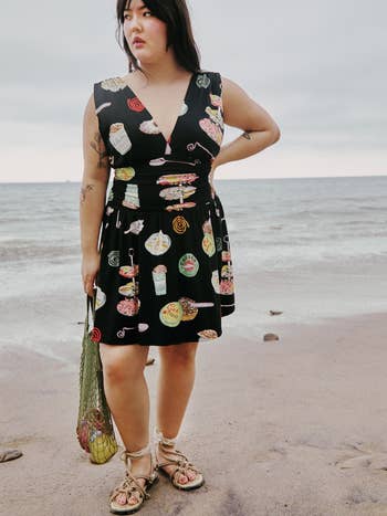 Person on a beach wearing a sleeveless dress with food prints, holding a net bag and wearing sandals
