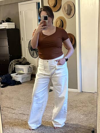 Person wearing a casual brownish  apical  and achromatic  wide-leg pants, taking a reflector  selfie successful  a chamber  with laundry baskets and hats successful  the background