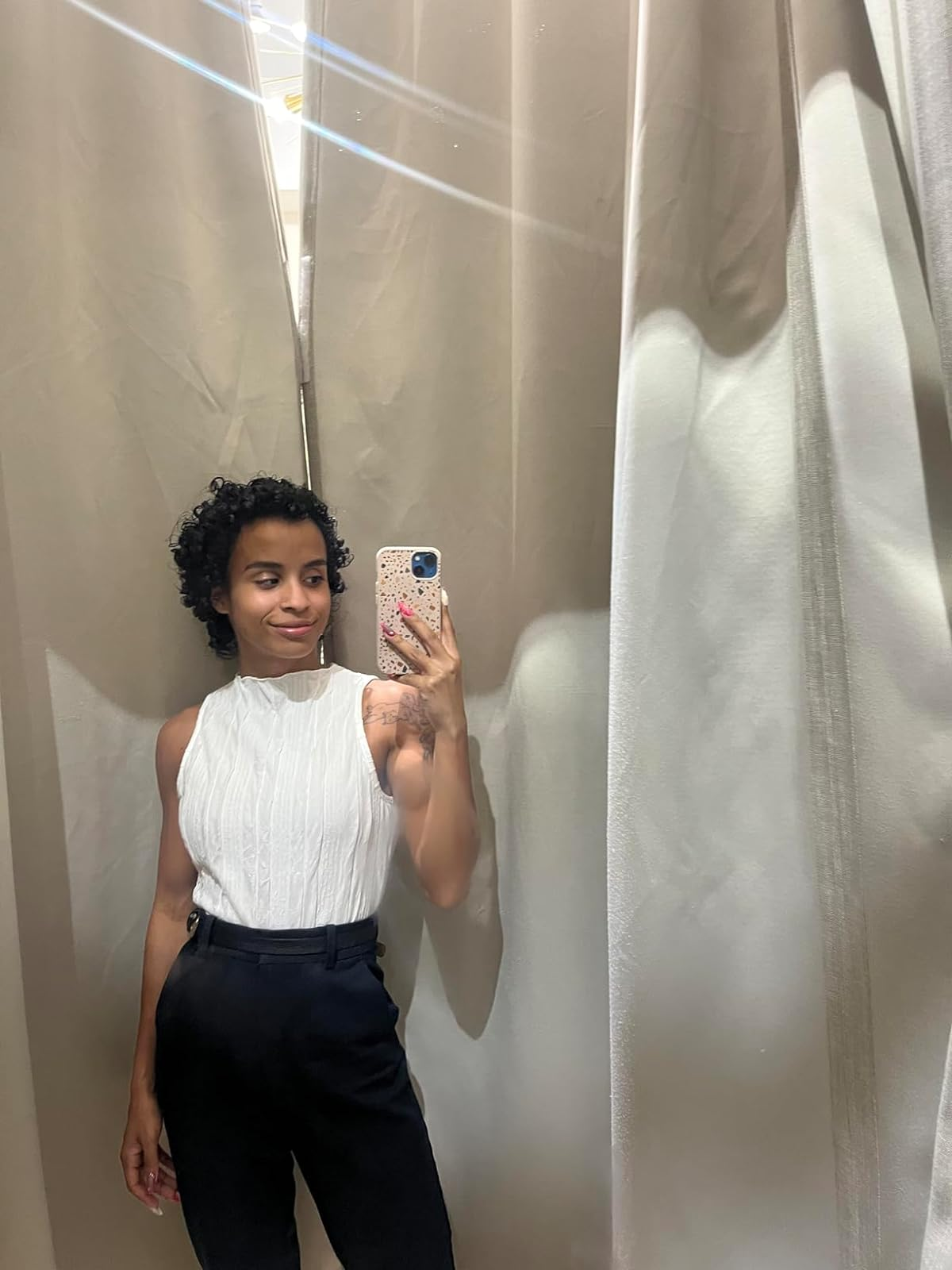 Person in fitting room trying on a white sleeveless top and black trousers, taking a mirror selfie