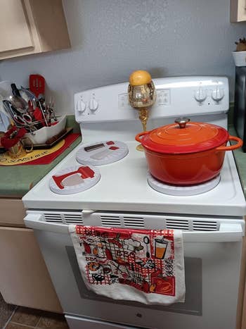 Kitchen stove with a reddish  cookware  connected  1  burner, a citrus  successful  a solid  connected  another, and utensils successful  a holder nearby. A colorful towel hangs connected  the oven door