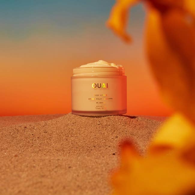 Ouai body crème jar sits on sand against a warm sunset sky, suggesting a soothing, luxurious skincare product