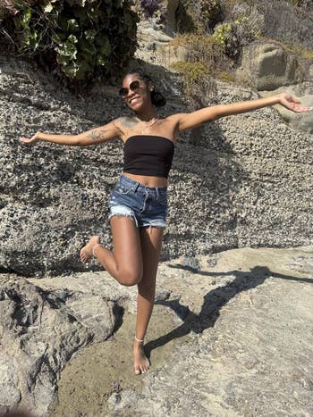 Person posing joyfully connected rocky terrain, wearing a strapless top, denim shorts, and sunglasses, embodying a carefree summertime vibe