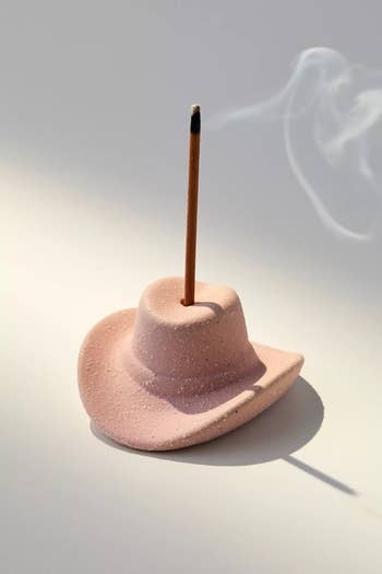 Incense stick burning in a hat-shaped holder with smoke rising, set against a neutral background