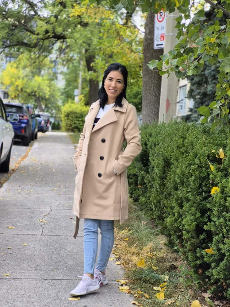Person successful  a beige trench coat, jeans, and sneakers stands connected  a tree-lined sidewalk, smiling astatine  the camera
