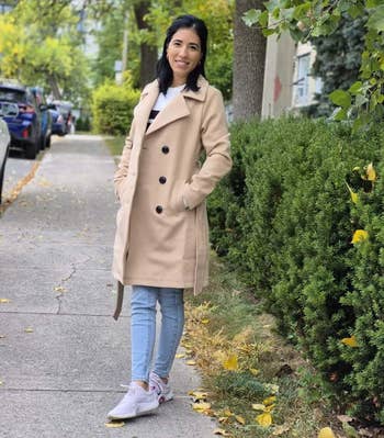 Person successful a beige trench coat, jeans, and sneakers stands connected a tree-lined sidewalk, smiling astatine the camera