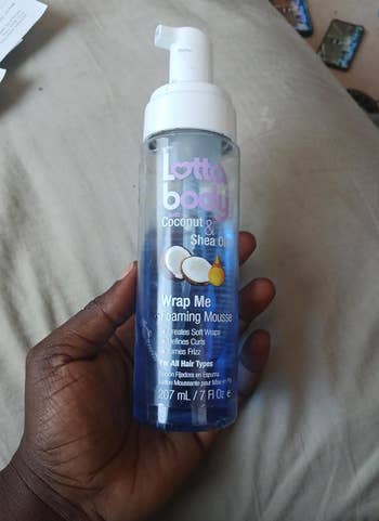 A hand holding a bottle of Lottabody Wrap Me Foaming Mousse with Coconut and Shea Oils, for all hair types, 207ml/7 fl oz