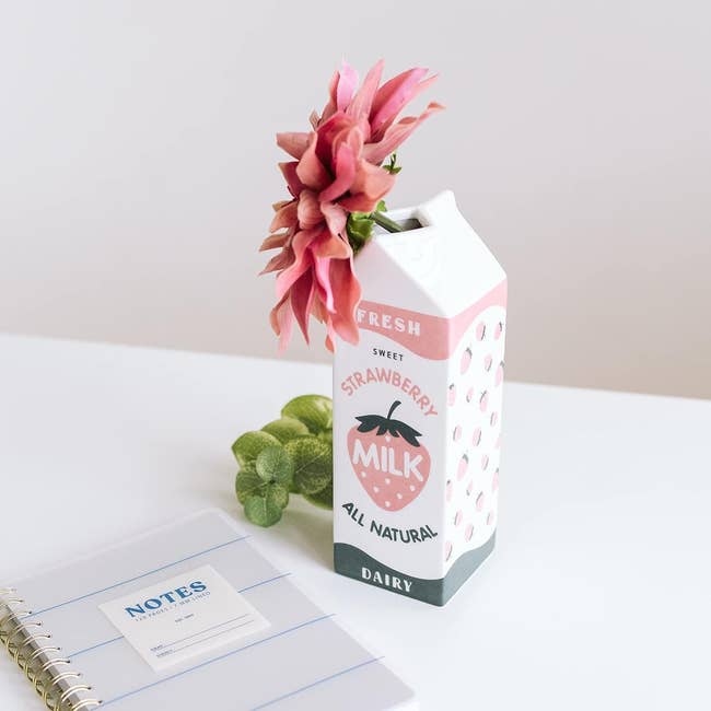 A white milk carton shaped vase with illustrated strawberry on it that 