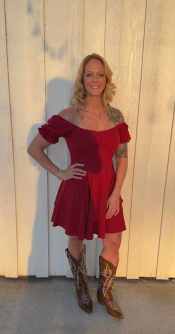 Person in an off-shoulder dress and cowboy boots poses by a textured wall, showcasing a casual yet chic look suitable for a shopping feature