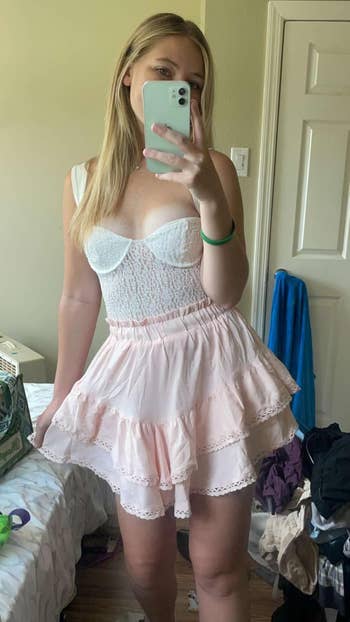 Person taking a selfie successful  a country   wearing a achromatic  lace apical  and pinkish  ruffled skirt. Bedroom items are disposable   successful  the background