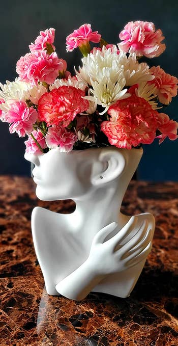 white vase shaped like the lower half of a face, neck, and shoulders with a hand with a floral arrangement sticking out