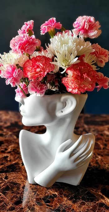 white vase shaped like the lower half of a face, neck, and shoulders with a hand with a floral arrangement sticking out