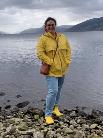 Person in a yellow jacket and jeans stands by a lake with mountains in the distance, carries a shoulder bag