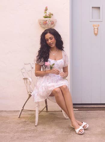 Model in a floral dress sitting outdoors, holding a pink flower, wearing sandals and a necklace. Elegant and casual look suitable for summer shopping